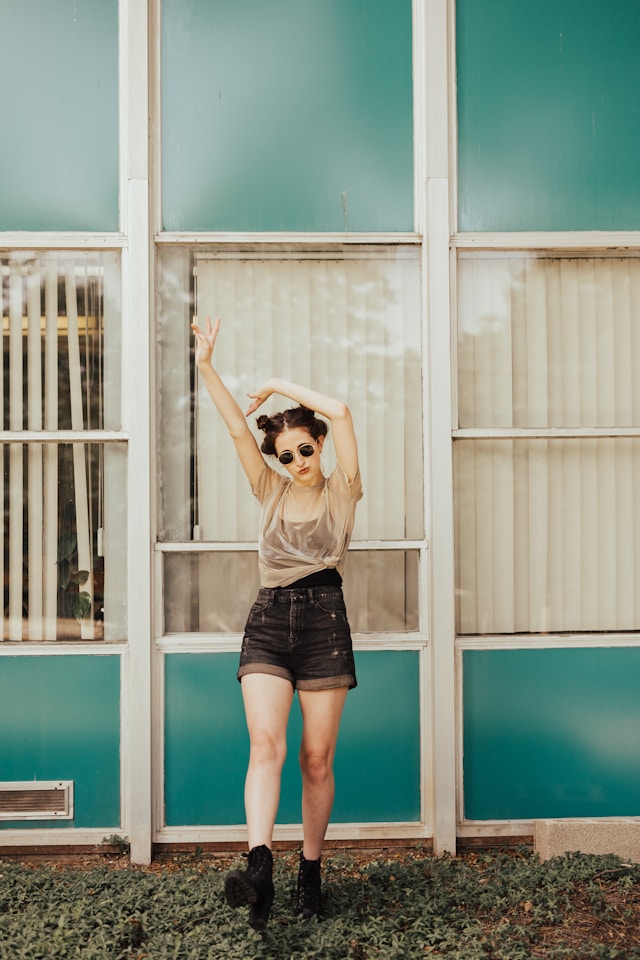 Woman in her early thirties stands in the sun with her hands in the air. She is wearing her hair in space buns and has sunglasses on. She is wearing boots, short denim shorts, and a black singlet, with a sheer nude t-shirt over the top. She is standing in front of a bright blue house with a row of windows in the middle of the frame.